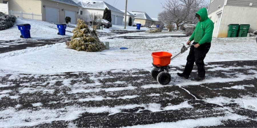 Worker spreading salt on a snow-covered residential driveway and sidewalk during winter to prevent ice buildup and ensure safe access.