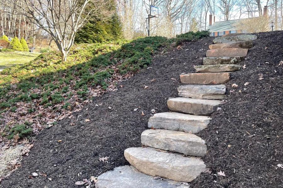 Rustic stone steps ascending a mulched hillside, bordered by green ground cover and trees, leading to a cozy residential property in a natural setting.