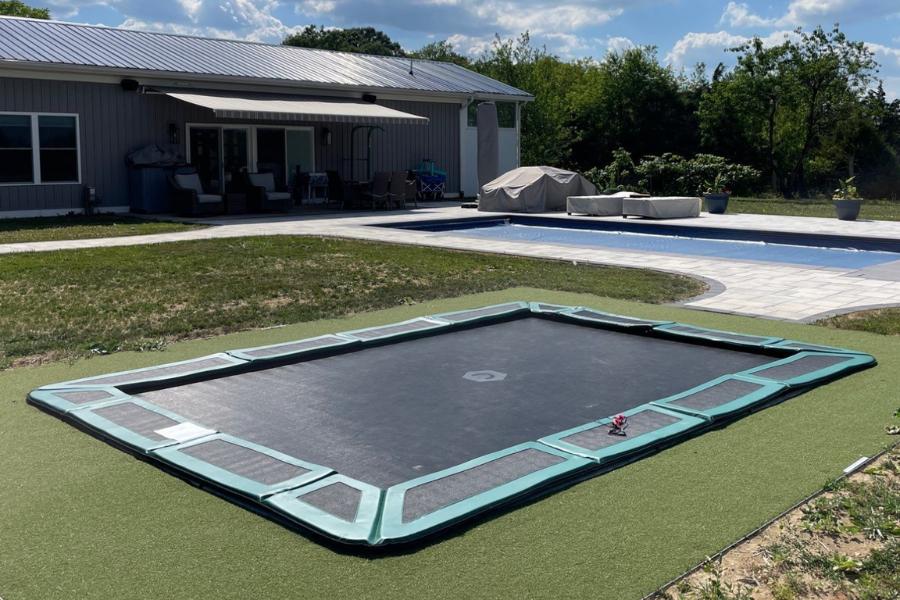 Backyard with an in-ground trampoline on artificial turf, adjacent to a patio and swimming pool, with a modern home and shaded seating area in the background.
