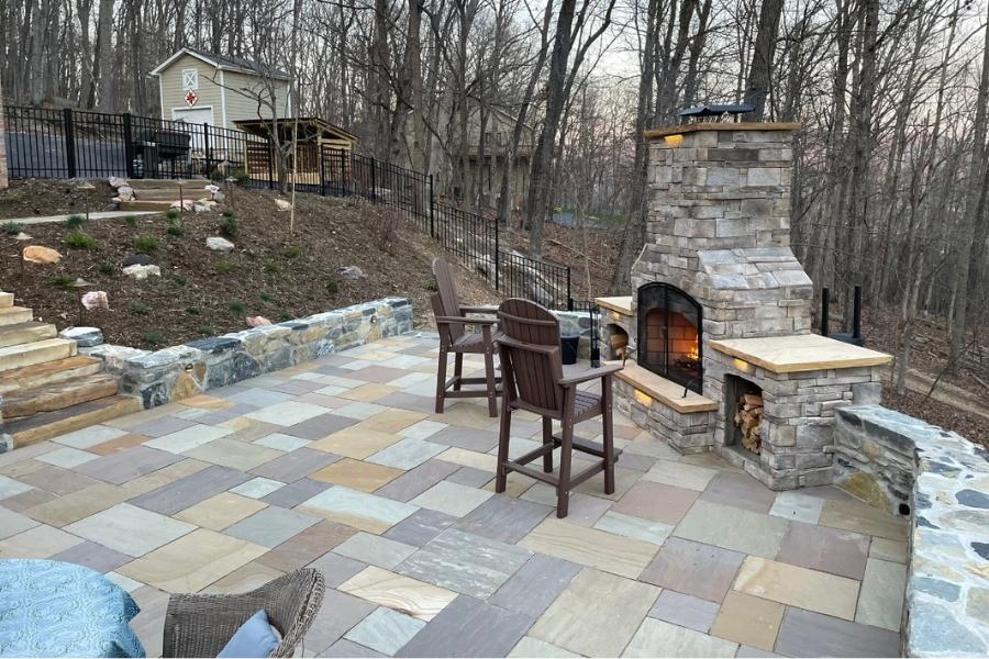 Cozy outdoor patio with a stone fireplace, surrounded by natural stone seating walls, overlooking a wooded backyard and hillside landscaping.