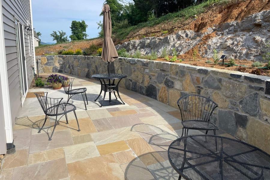 Outdoor patio with stone pavers, wrought iron tables and chairs, bordered by a decorative stone retaining wall and hillside landscaping.