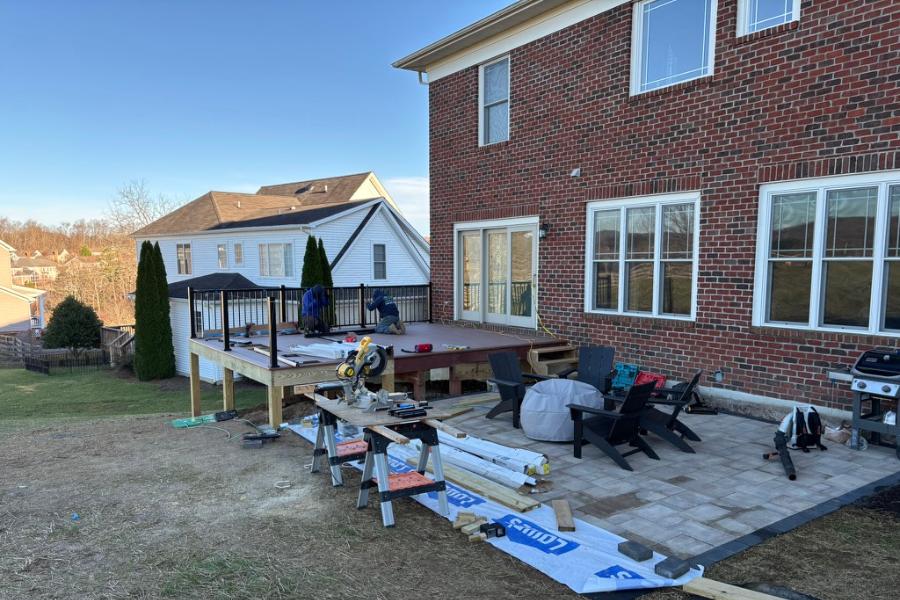 Backyard construction project featuring a raised deck under construction, adjacent to a completed patio with pavers and outdoor seating, attached to a brick house.