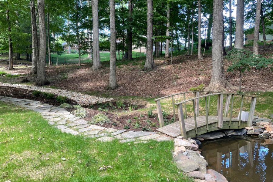 Scenic backyard landscape featuring a small wooden bridge over a pond, a stone pathway, and a dry creek bed surrounded by tall trees and lush greenery.