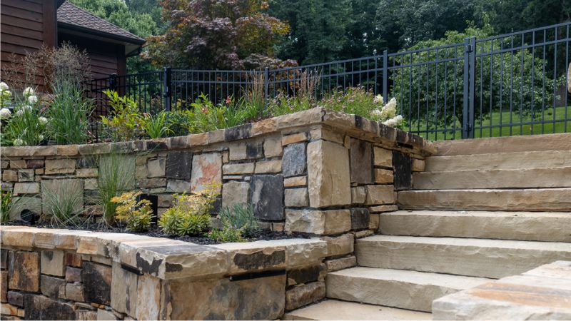 Elegant stone retaining wall with integrated planters, natural stone steps, and a black metal fence, enhancing a landscaped backyard.