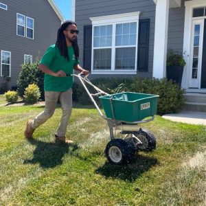 Professional landscaper applying lawn treatment using a Lesco broadcast spreader on a green lawn in front of a modern suburban home.