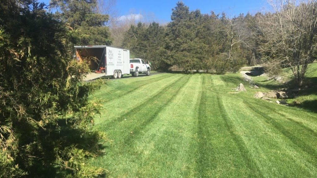 Freshly mowed lawn with professional striping, surrounded by trees, with a landscaping trailer and truck parked nearby for lawn care services.